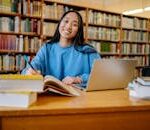Smiling student studying in a library with books and laptop, showcasing education and focus.